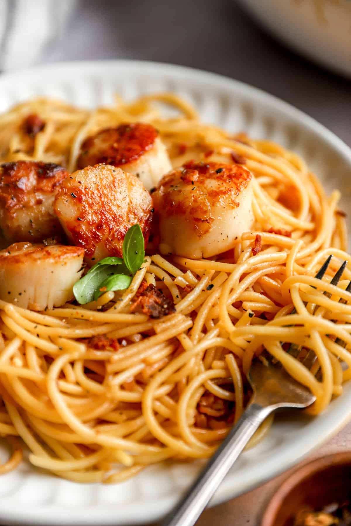 closeup of a fork swirled in a plate of pasta topped with pan-seared scallops from the side.