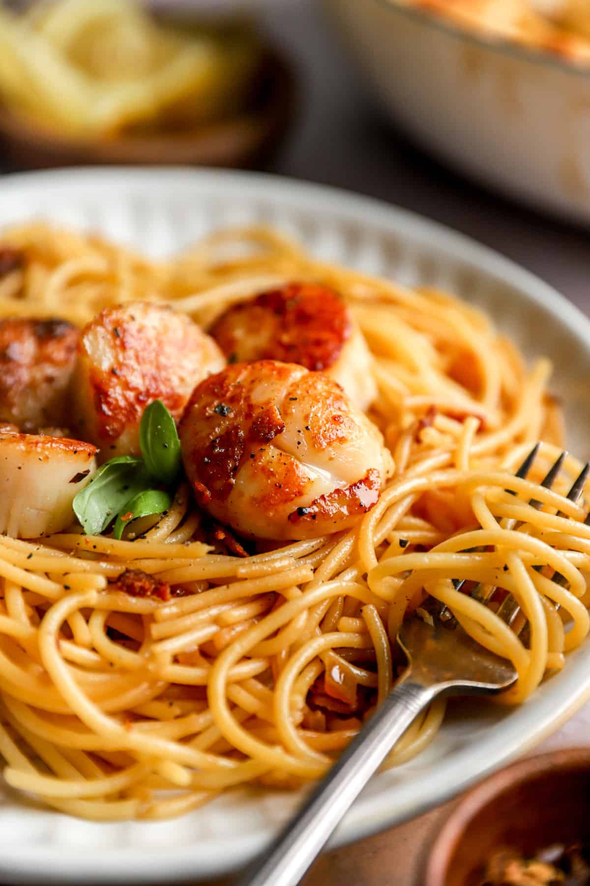 closeup of a fork swirled in a plate of pasta topped with pan-seared scallops from the side.