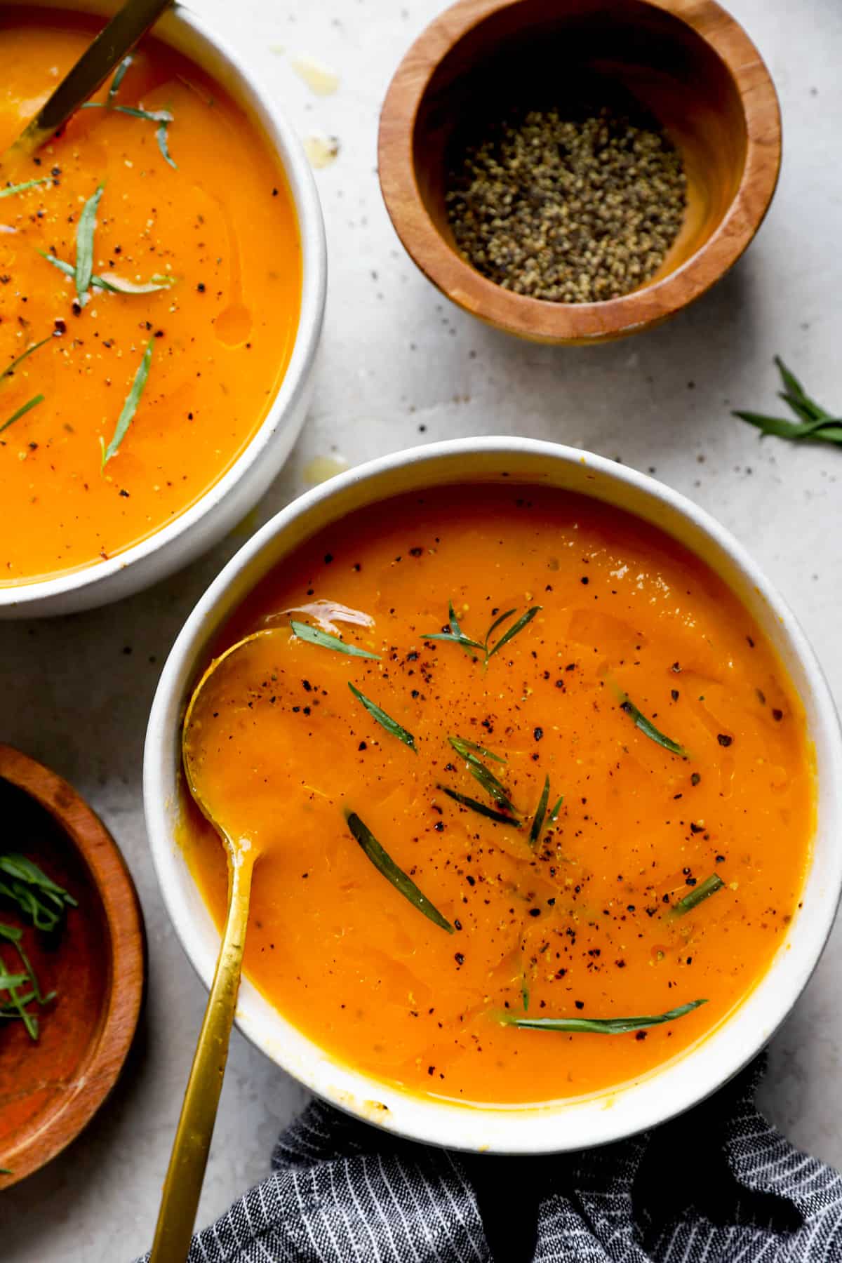 closeup image of a bowl of squash soup with another bowl of soup and two bowls of fresh tarragon next to it.
