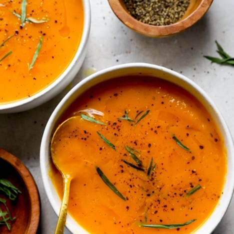 closeup image of a bowl of squash soup with another bowl of soup and two bowls of fresh tarragon next to it.