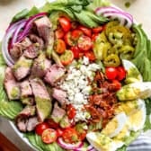 an entire steak cobb salad in a white bowl - think a bed of lettuce, diced cherry tomatoes, diced red onions, sliced hard boiled egg, chopped bacon, sliced steak.