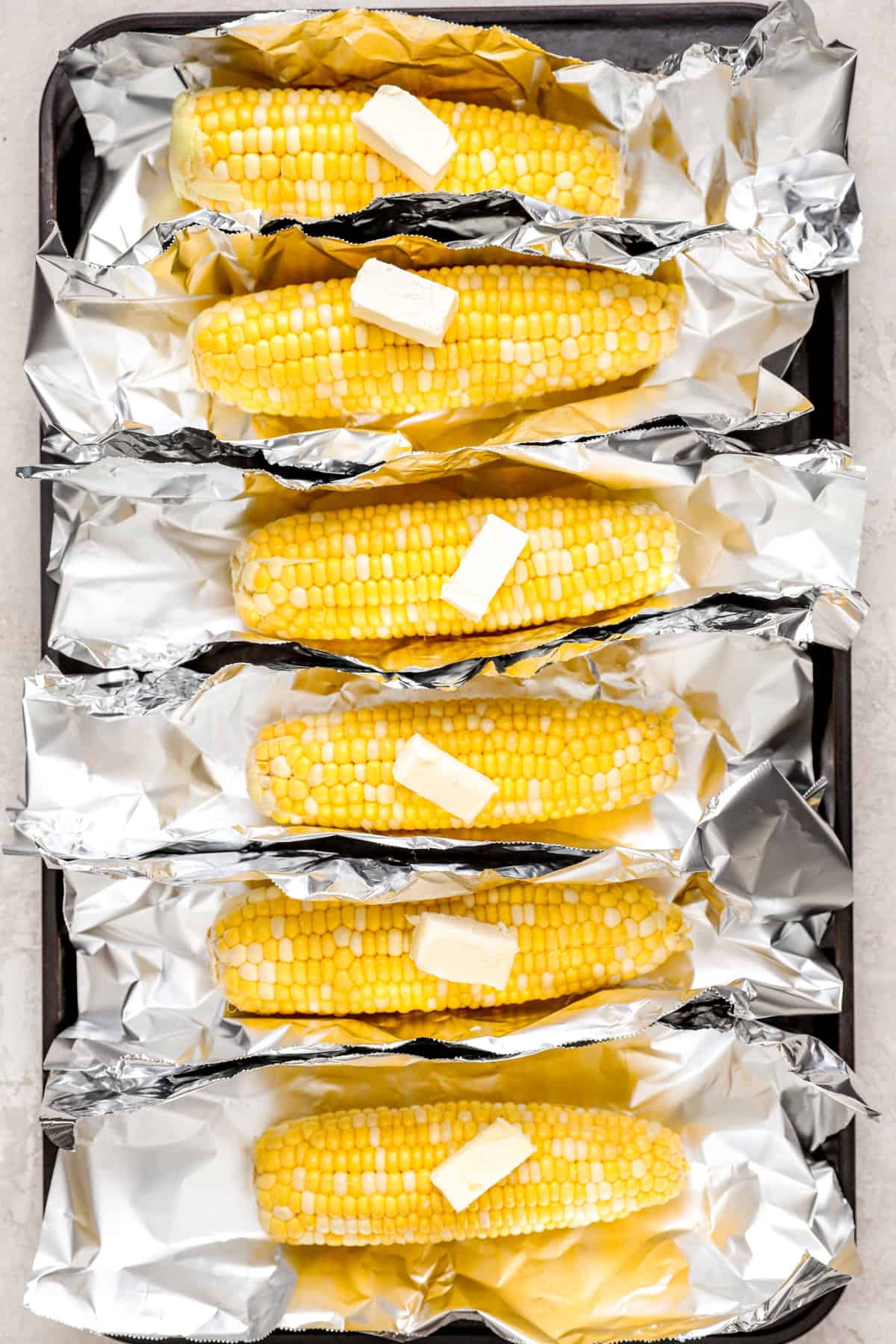 cobs of corn lined on a baking sheet with foil and topped with butter.