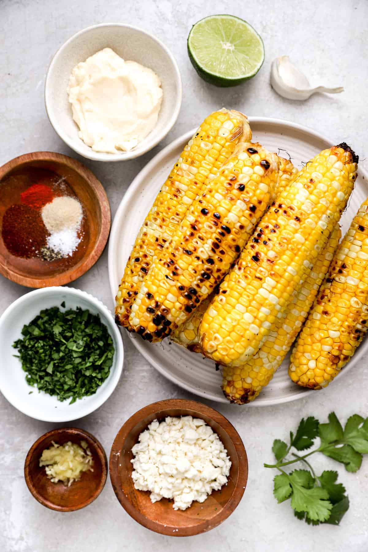 above image of grilled corn on the cob piled on a plate surrounded by street corn salad ingredients.