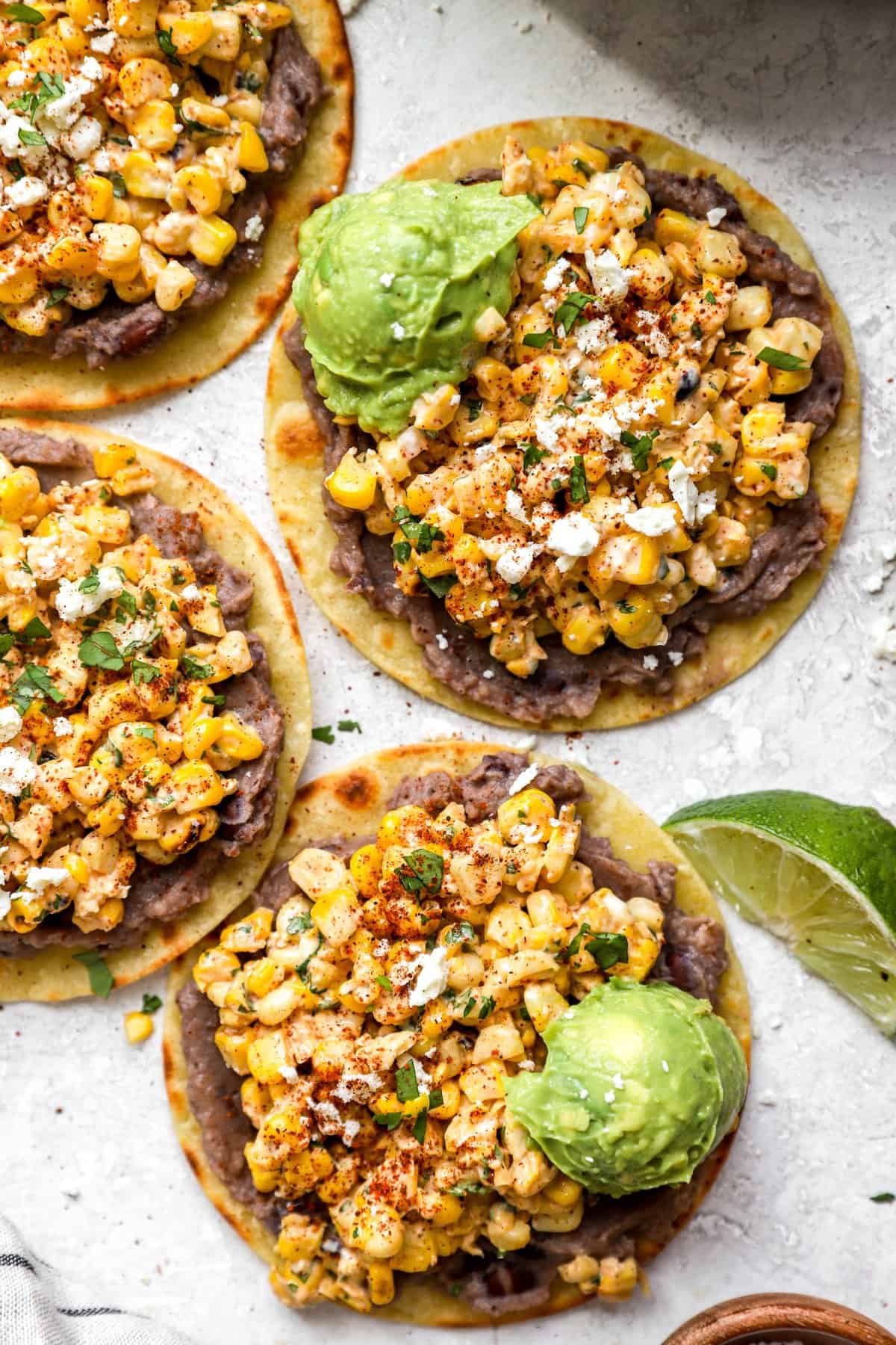 three tostadas topped with Mexican street corn dip and garnished with lime.