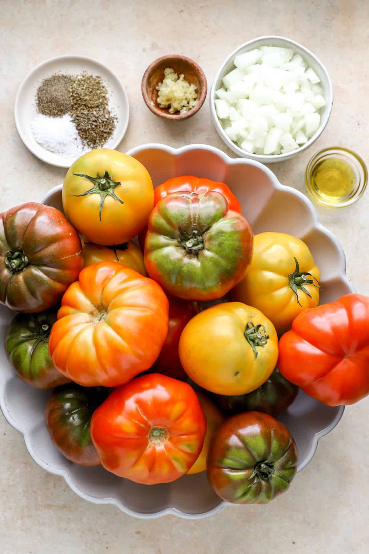 A white bowl full of colorful, ripe tomatoes and a smaller bowl of chopped onions, a small plate of spices, and two small condiment bowls with oil and garlic.