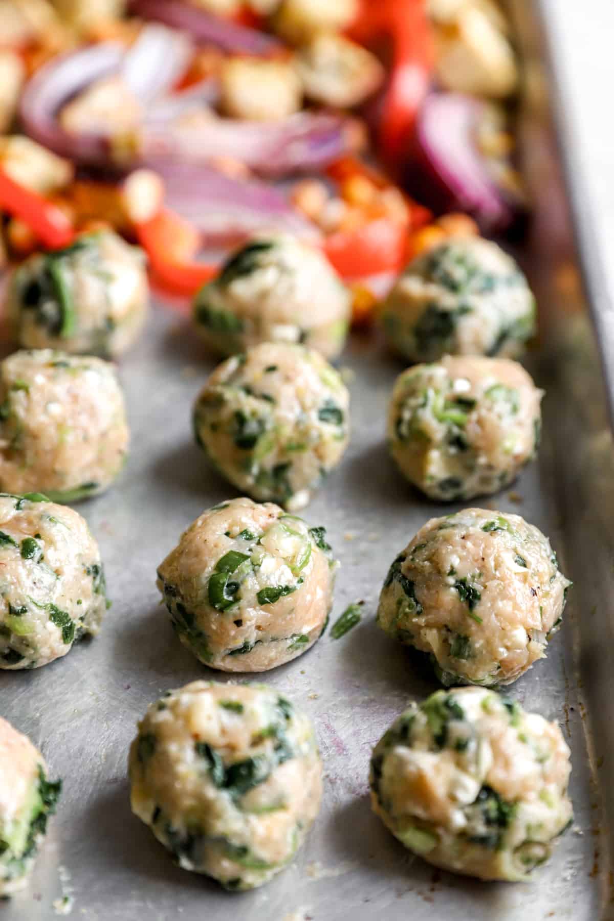 unbaked chicken meatballs on a baking sheet with veggies from the side.