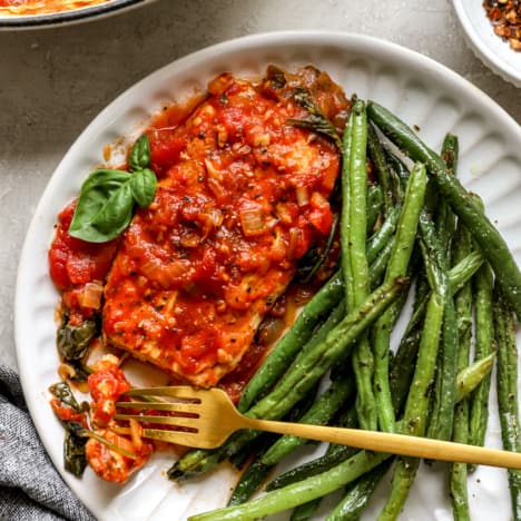 above image of a chicken in tomato sauce on a plate with roasted green beans and a gold fork.