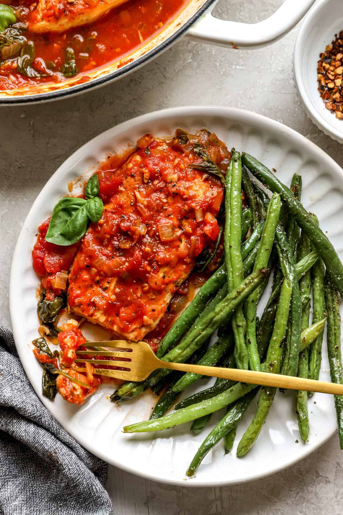 above image of a chicken in tomato sauce on a plate with roasted green beans and a gold fork. 