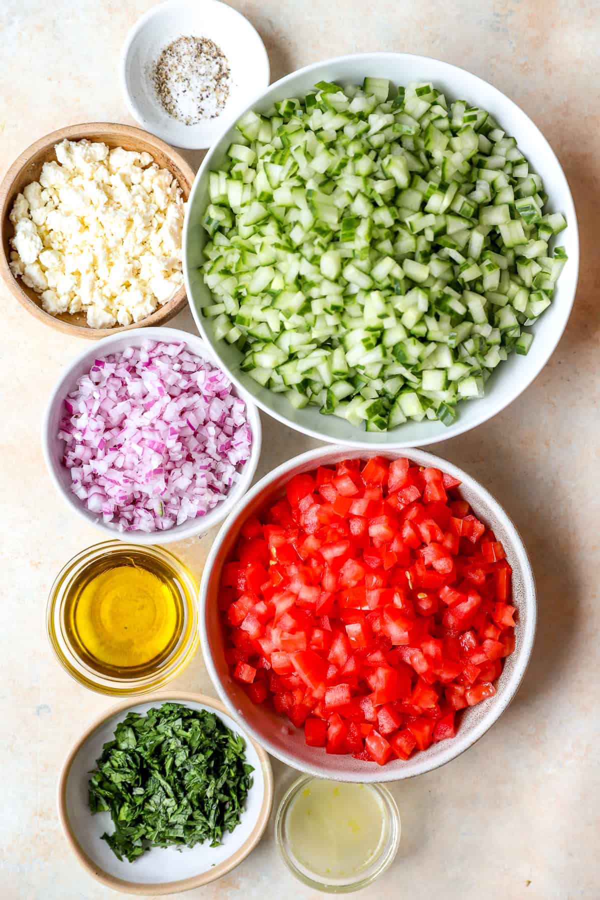 Multiple bowls filled with finally chopped cucumber, red onion, tomato, feta, parsley and olive oil with lemon juice.