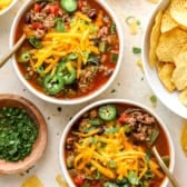 Two bowls of taco soup topped with favorite toppings and showing a bowl of chips and fresh jalapeños on the side.