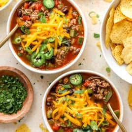 Two bowls of taco soup topped with favorite toppings and showing a bowl of chips and fresh jalapeños on the side.