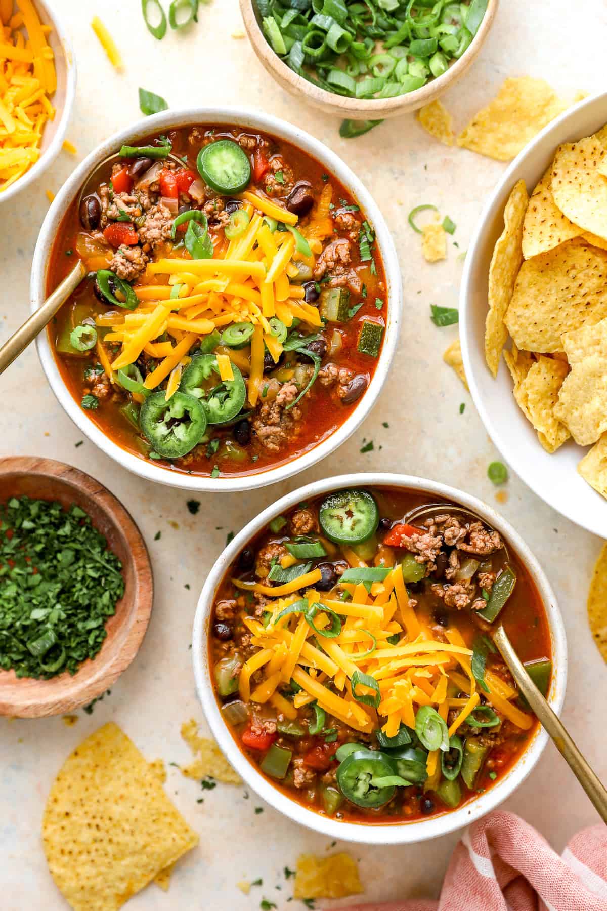 Two bowls of taco soup topped with favorite toppings and showing a bowl of chips and fresh jalapeños on the side.