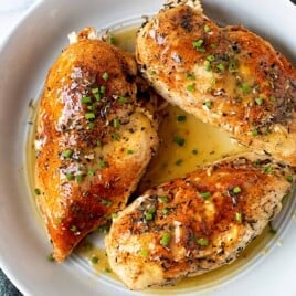 Fully cooked chicken breasts on a plate with pan sauce and herbs.
