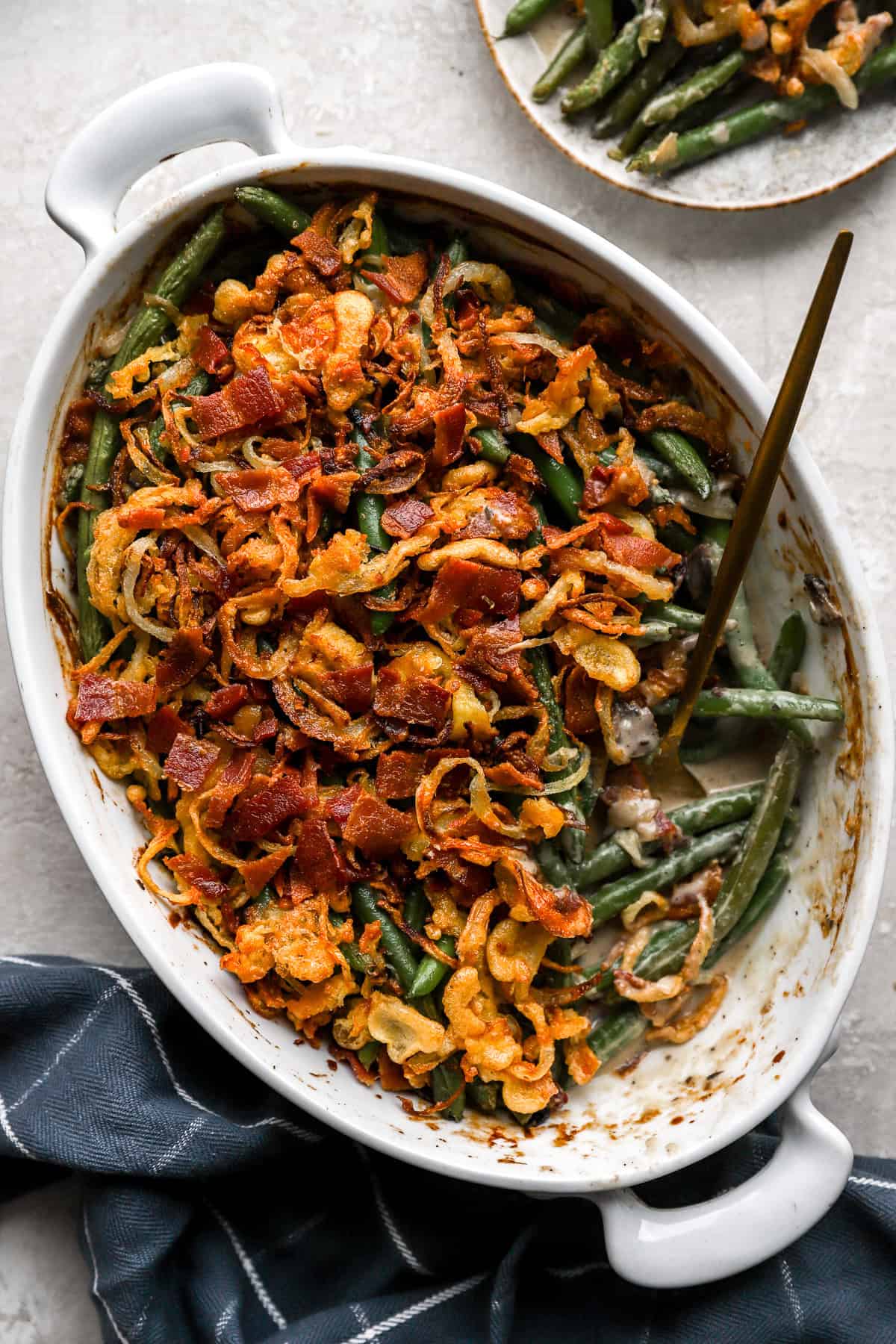 Fully cooked green bean casserole in a white baking dish with a blue napkin underneath and a spoon for serving.