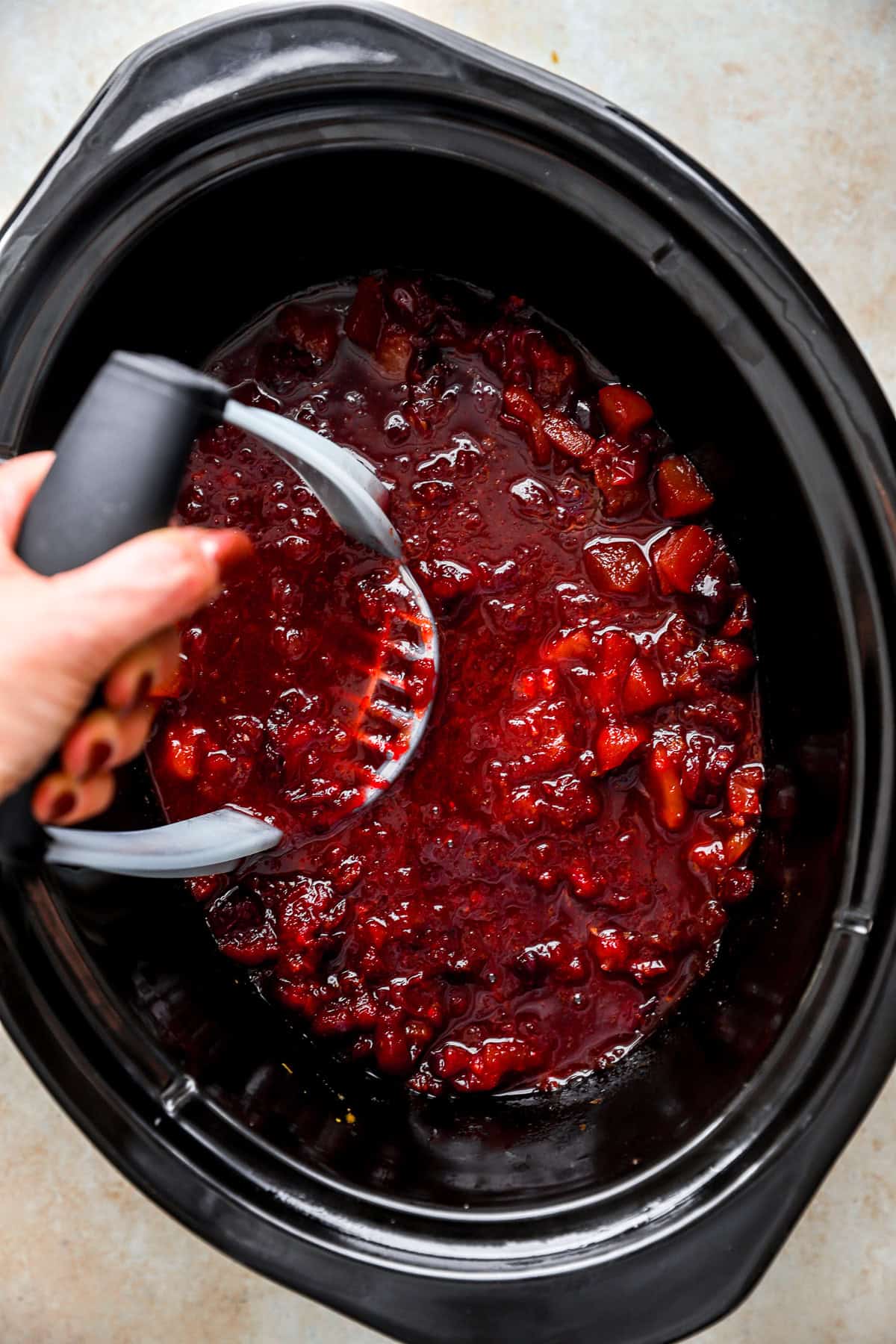 Black crockpot filled with mashed up cranberry sauce with a hand holding a potato masher.