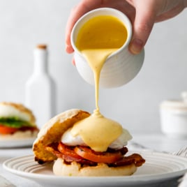 hollandaise sauce being drizzled over an eggs benedict breakfast sandwich on a plate.