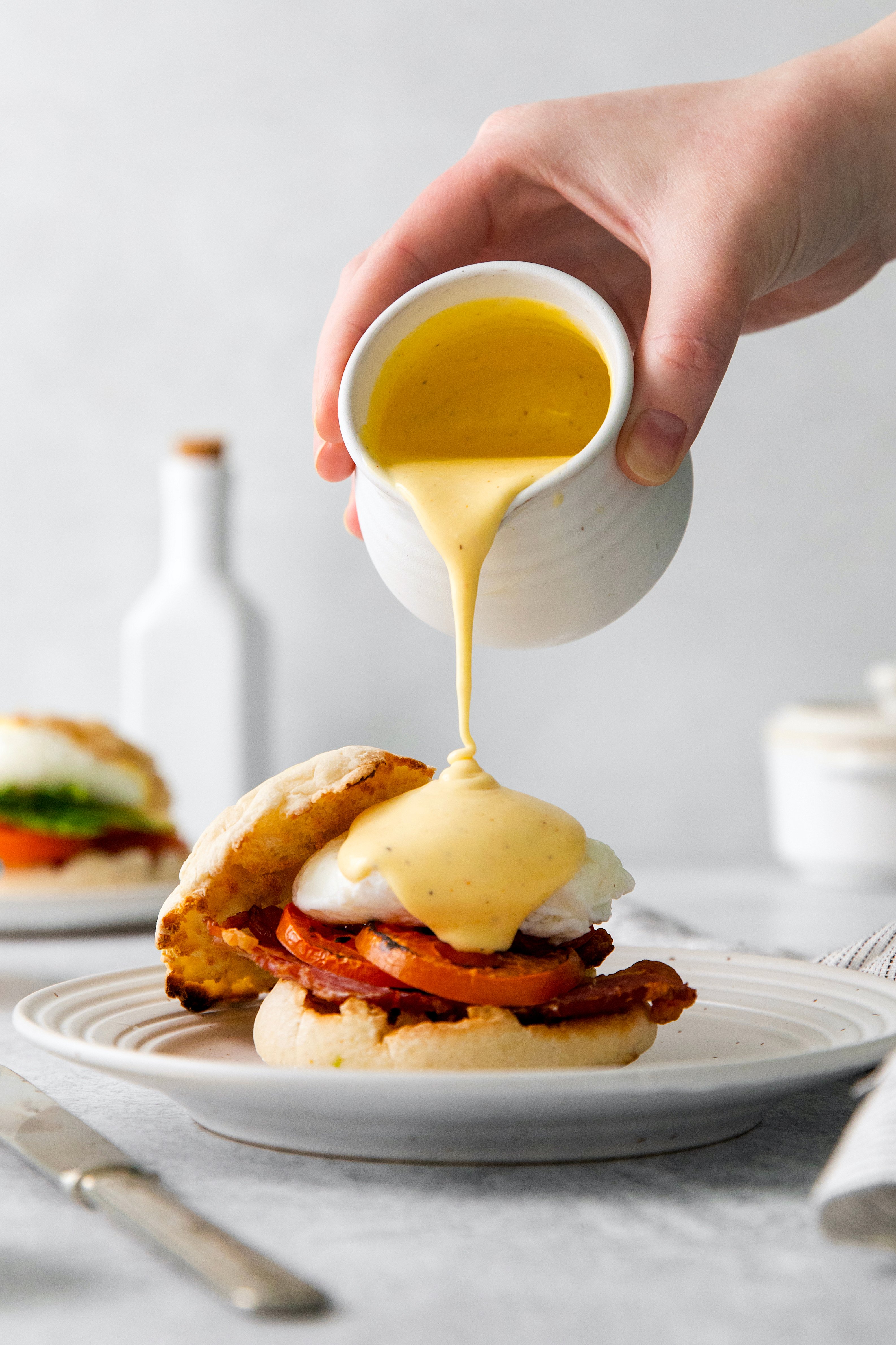 hollandaise sauce being drizzled over an eggs benedict breakfast sandwich on a plate.