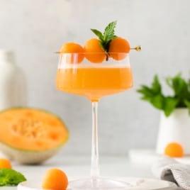 side view of a glass with a cantaloupe ginger cocktail garnished with melon balls and mint.