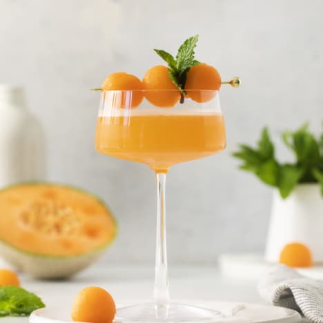 side view of a glass with a cantaloupe ginger cocktail garnished with melon balls and mint.