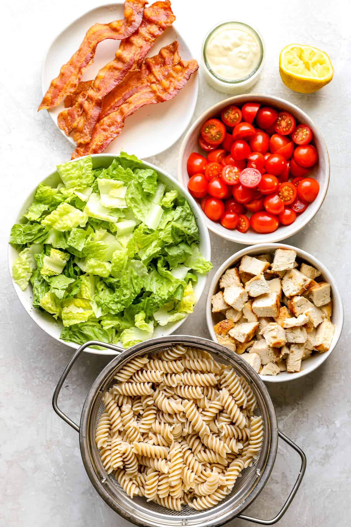 White bowls filled with ingredients. Lettuce in one, tomatoes in one, grilled chicken in one and crispy bacon in one with cooked pasta in a silver colander.