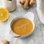 slightly side view overhead of a bowl of dressing next to fresh ginger and half of an orange.