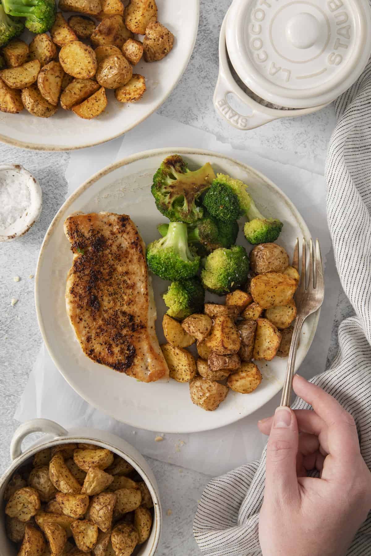 a fork being placed onto a plate with roasted potatoes, broccoli, and meat.