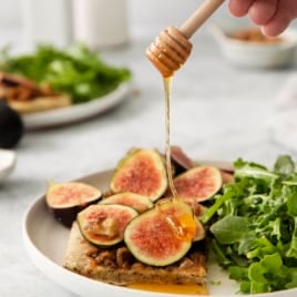 honey being drizzled over a slice of roasted fig pizza on a plate with an arugula salad.