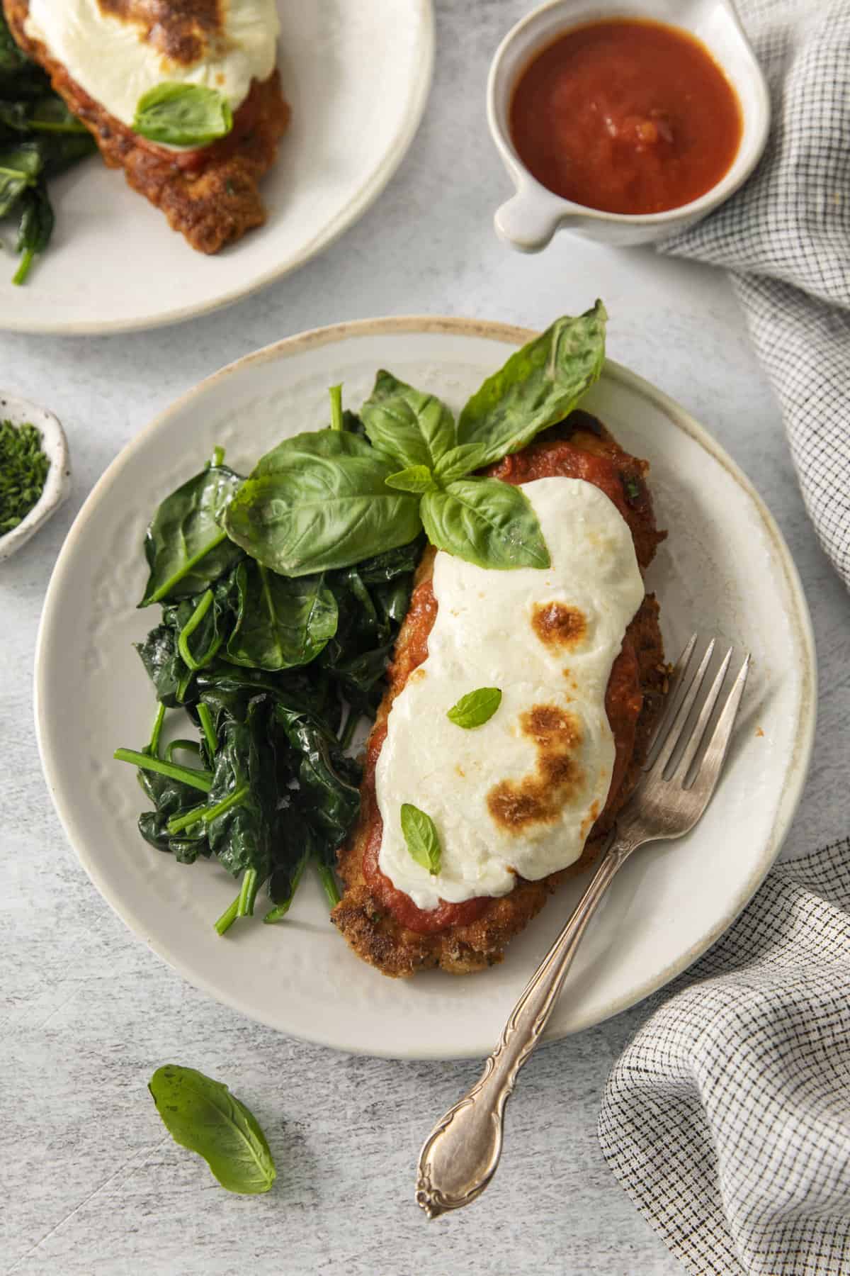 above image of a piece of chicken parmesan topped with fresh basil with a side of sautéed greens.