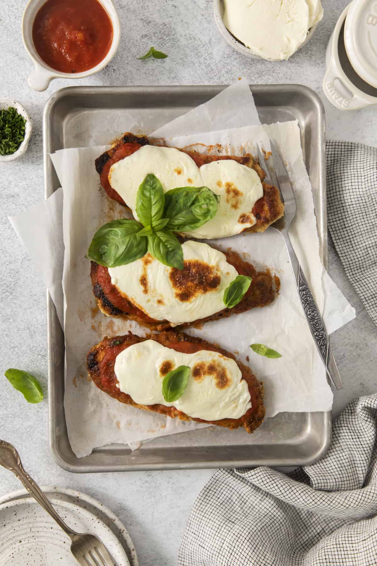 three pieces of chicken parmesan on a baking sheet from above garnished with fresh basil leaves.