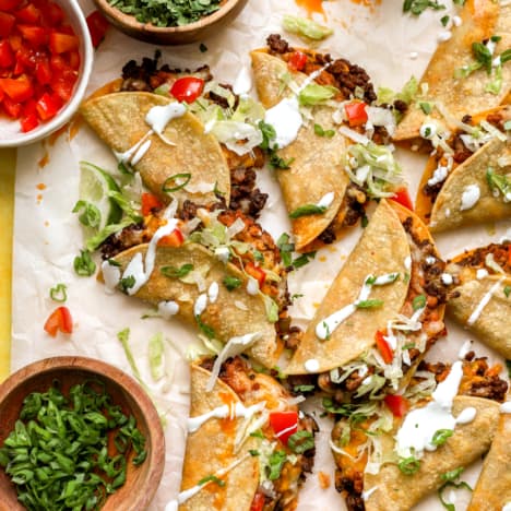a pile of ground beef tacos on a piece of parchment paper surrounded by bowls of toppings.