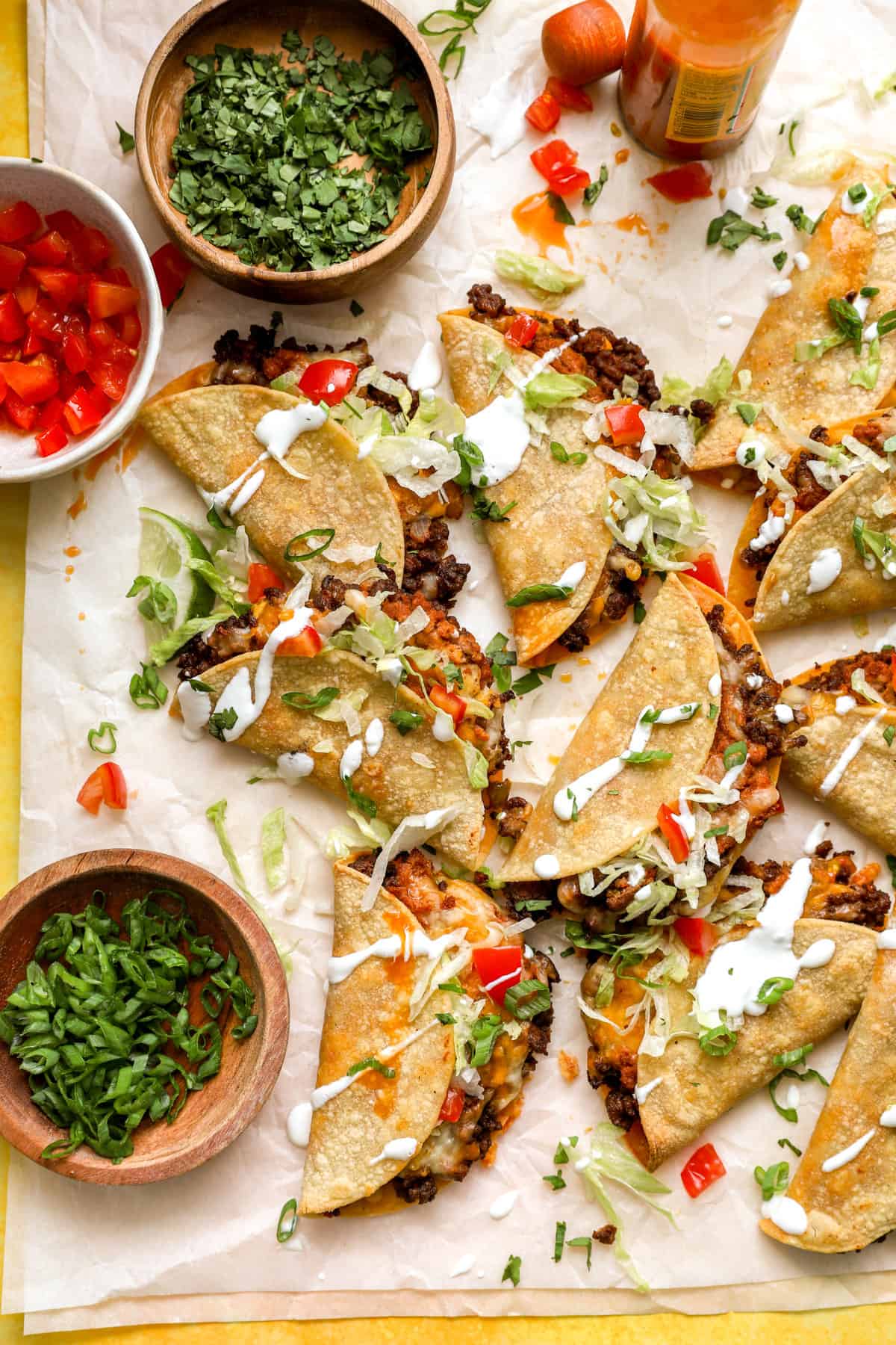 a pile of ground beef tacos on a piece of parchment paper surrounded by bowls of toppings. 