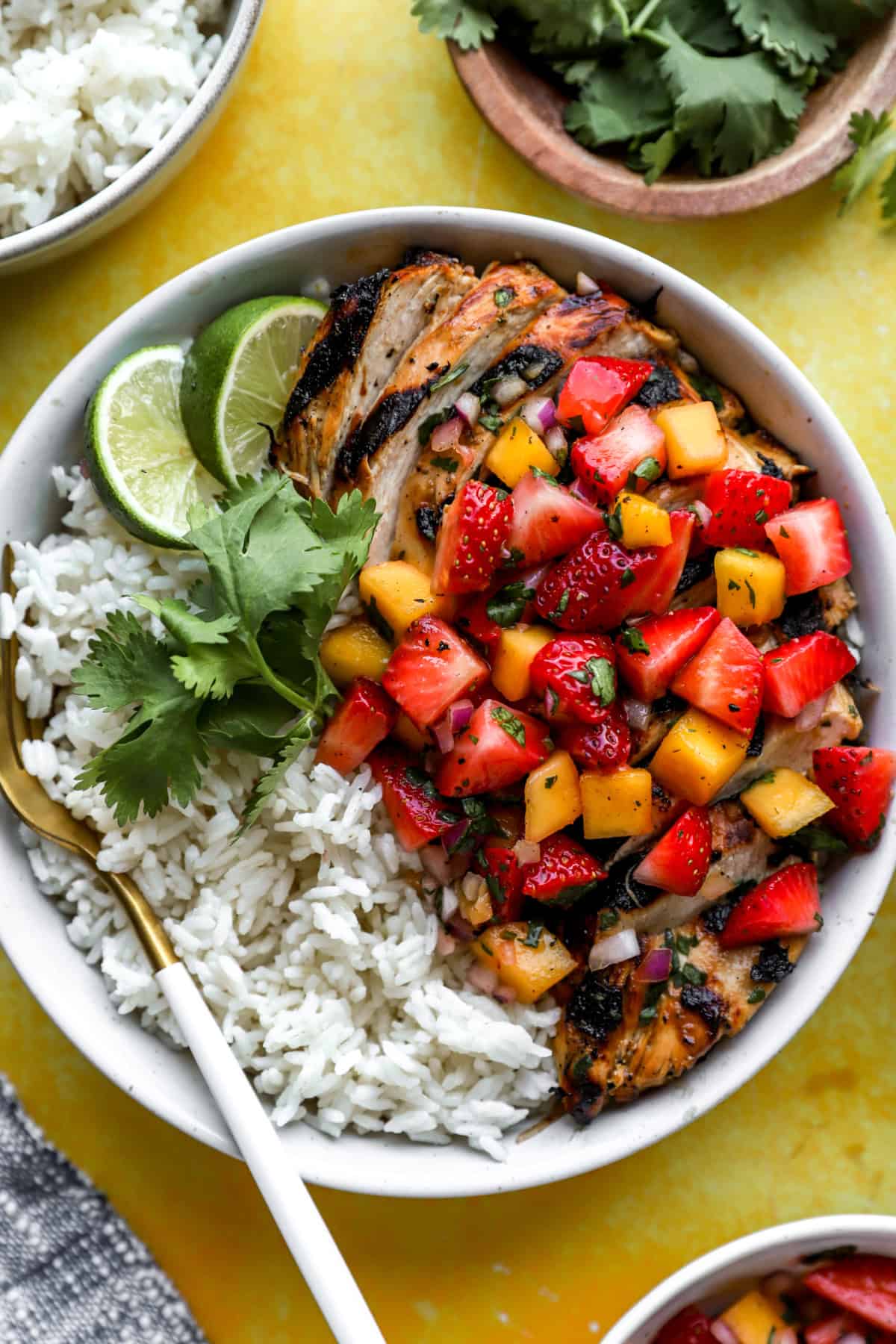 A rice bowl with grilled chicken, strawberry mango salsa, lime wedges, and cilantro. 