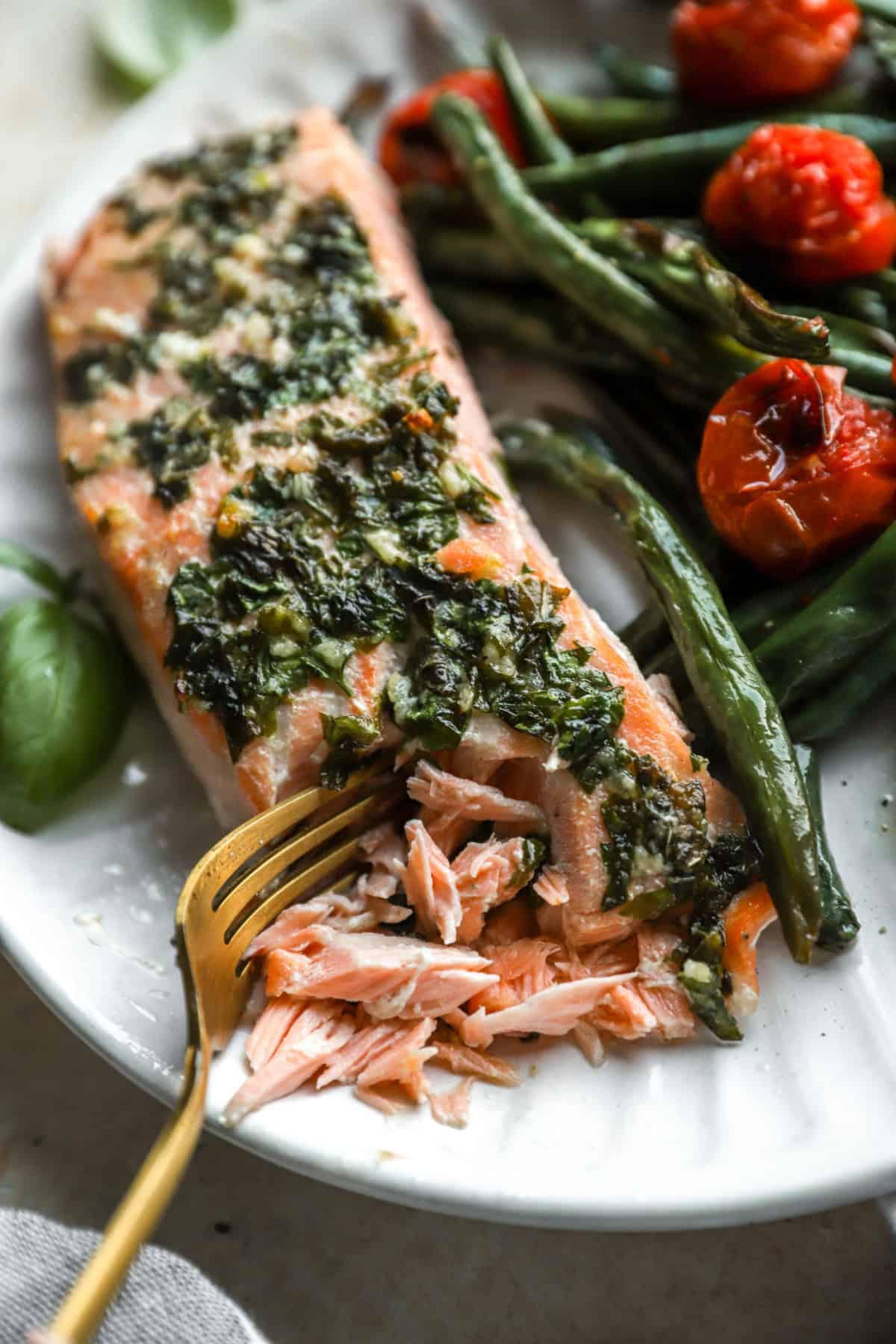 A fork flaking a bite of herb butter salmon on a plate with tomatoes and green beans. 
