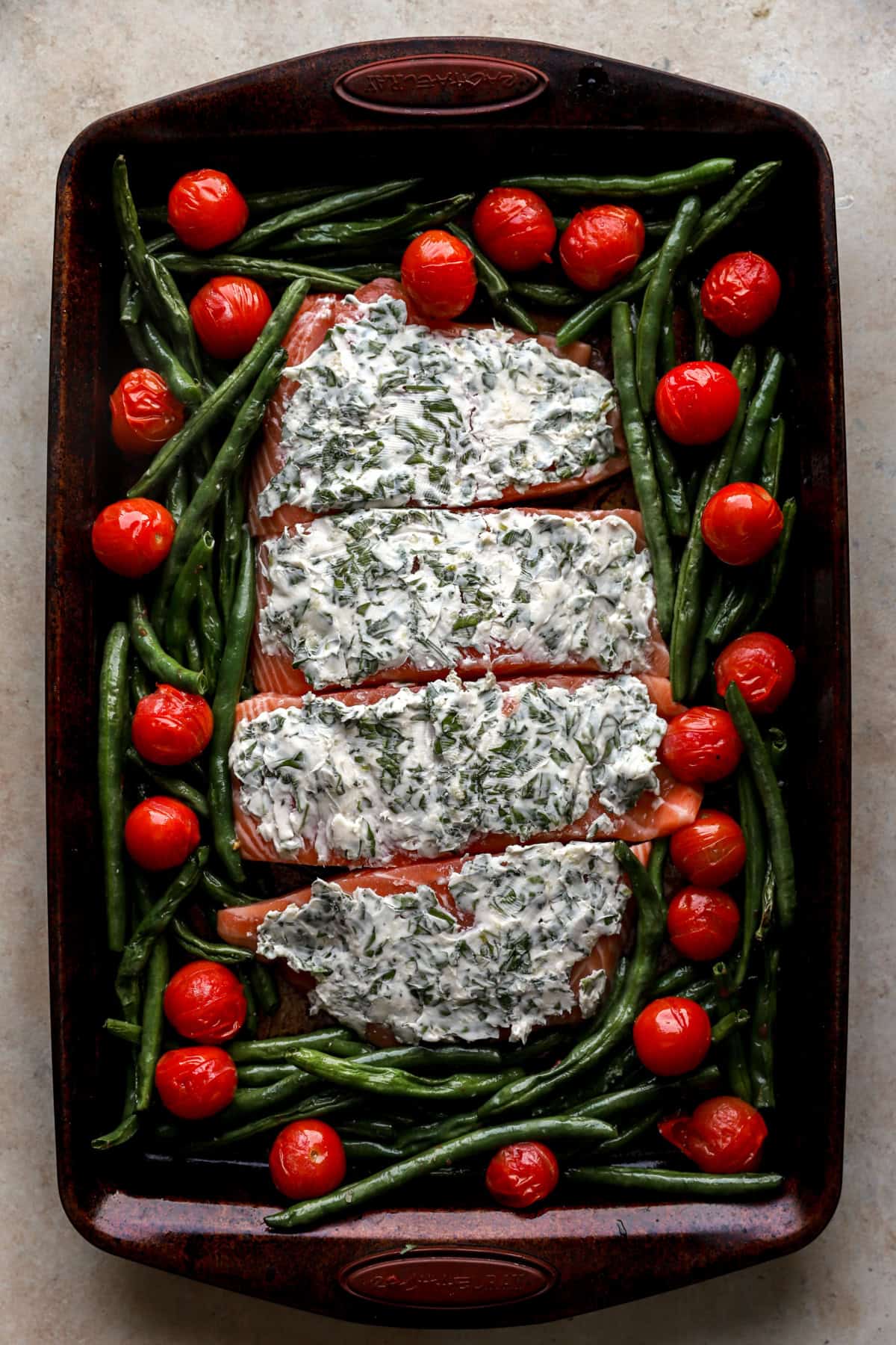Raw herb butter salmon filets on a baking sheet with tomatoes and green beans. 
