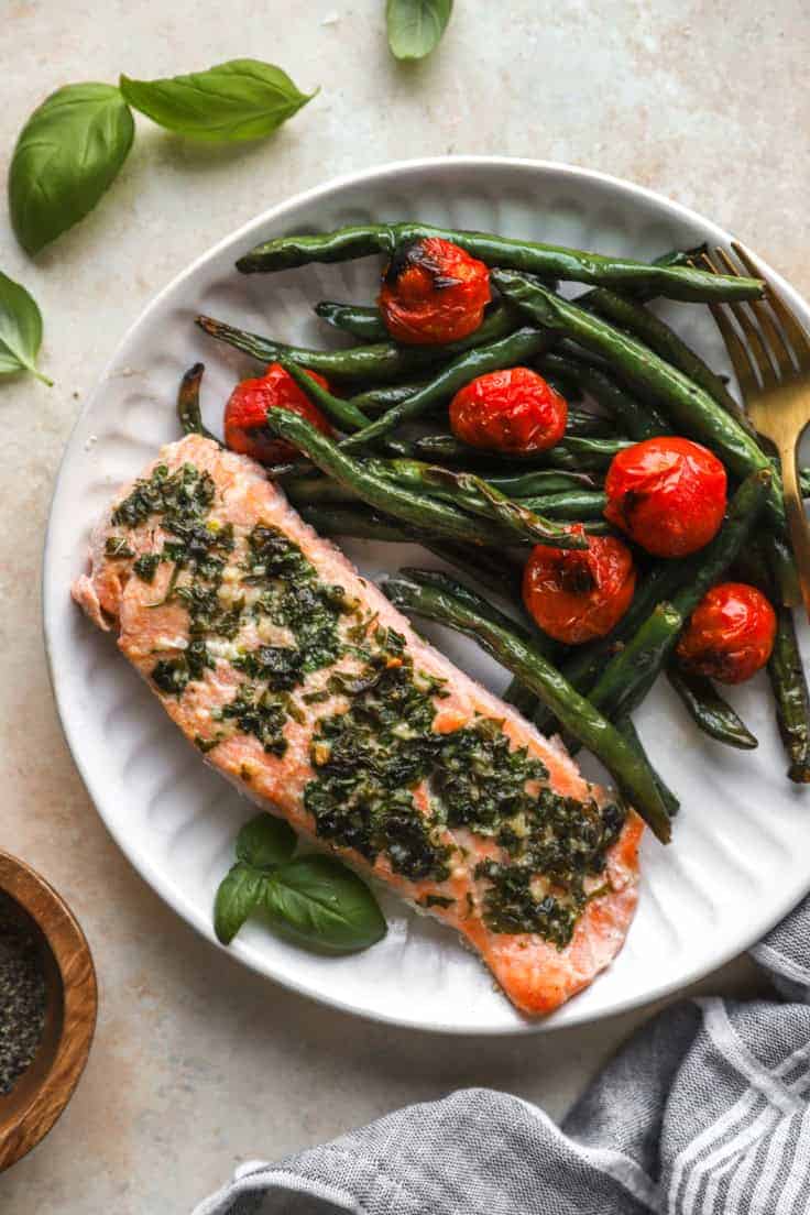 a white plate with a piece of herb butter salmon and roasted green beans with cherry tomatoes on the side with a fork. 