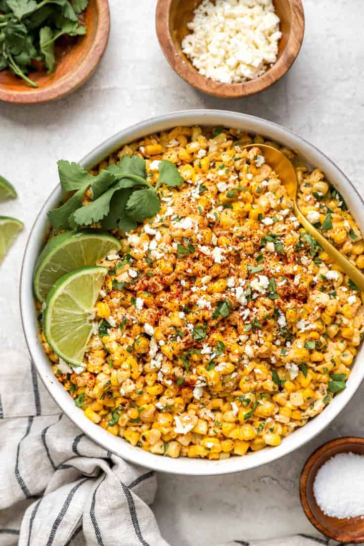 Bowl of roasted street corn with two limes wedges, fresh cilantro and a gold spoon.
