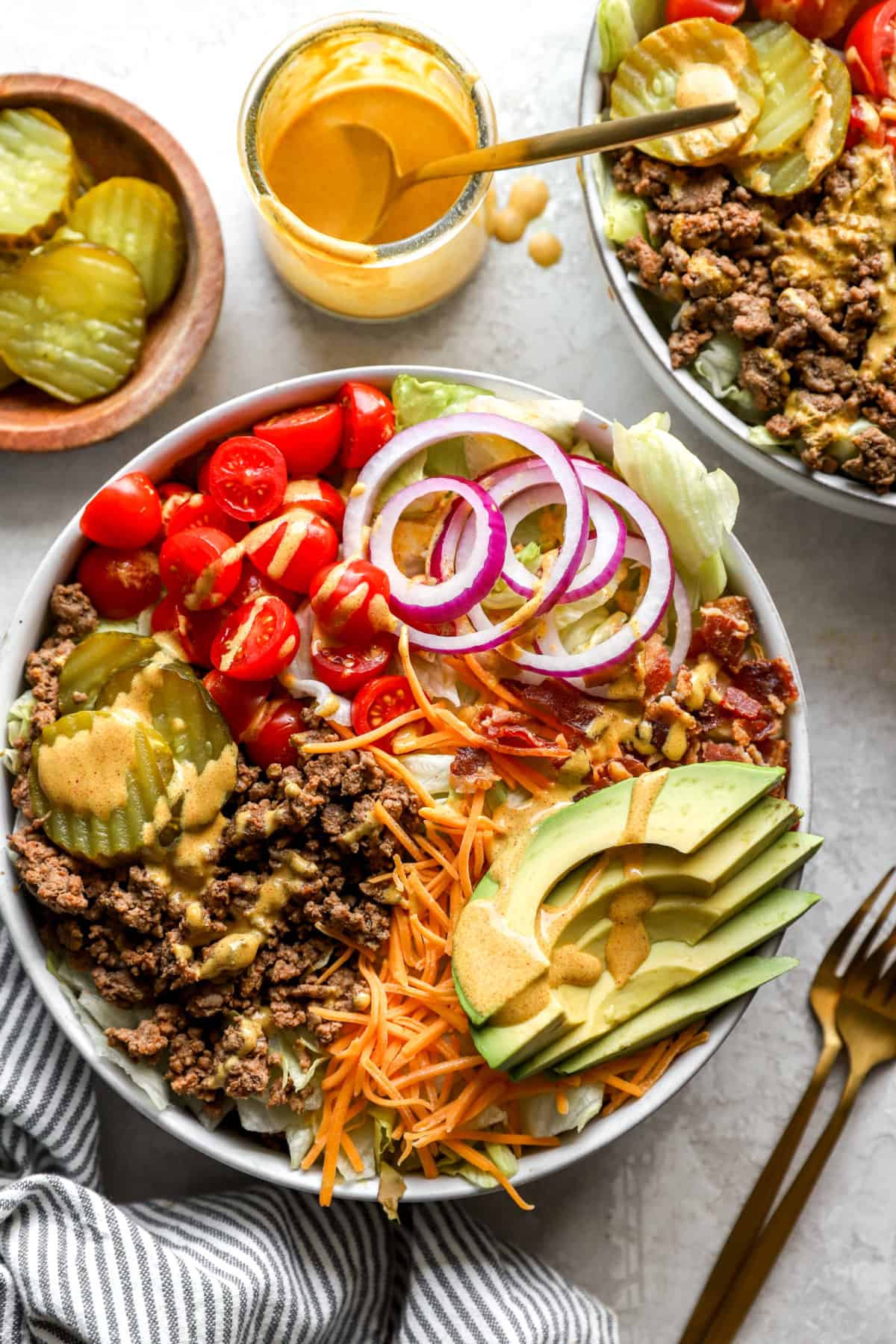 A bowl of cheeseburger salad surrounded by bowls of toppings. 