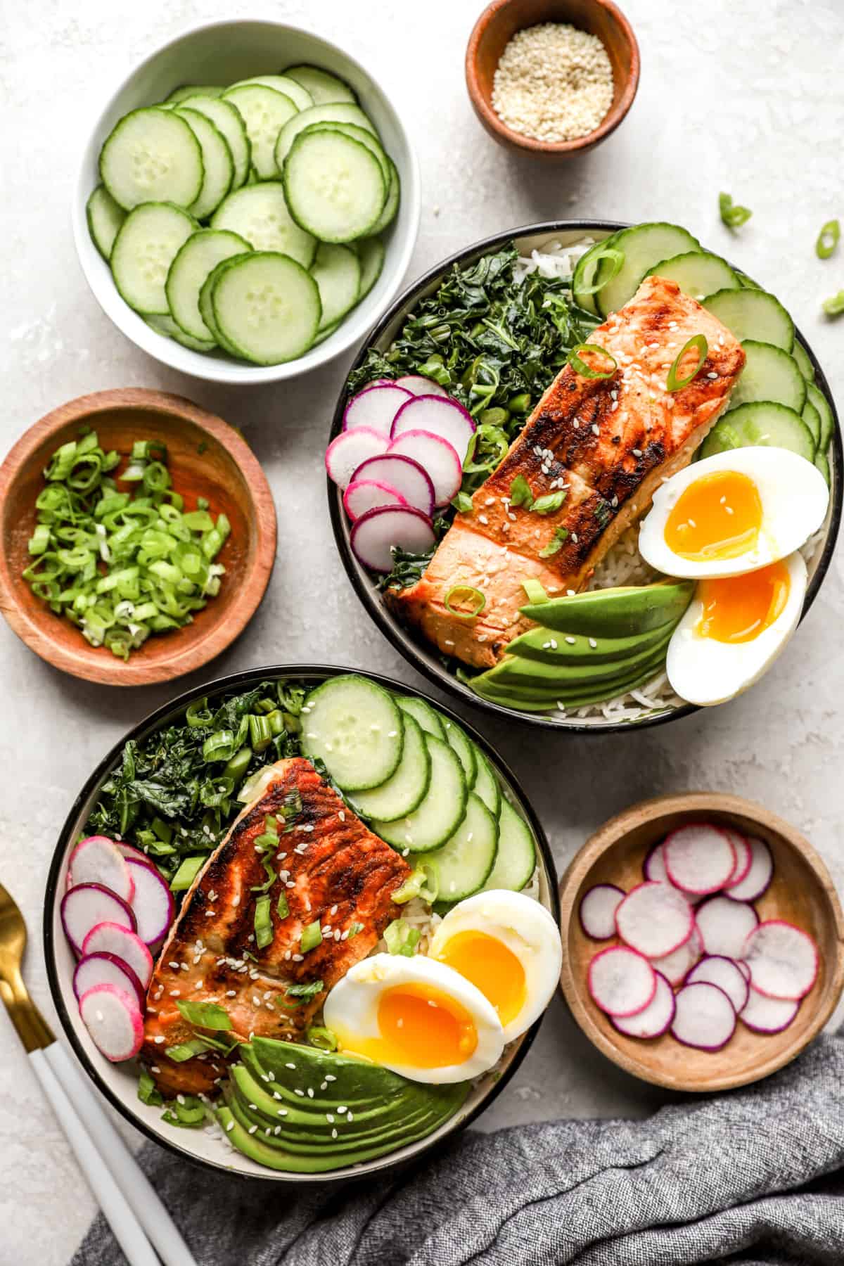 Overhead image of two grilled salmon bowls with bowls of toppings all around.