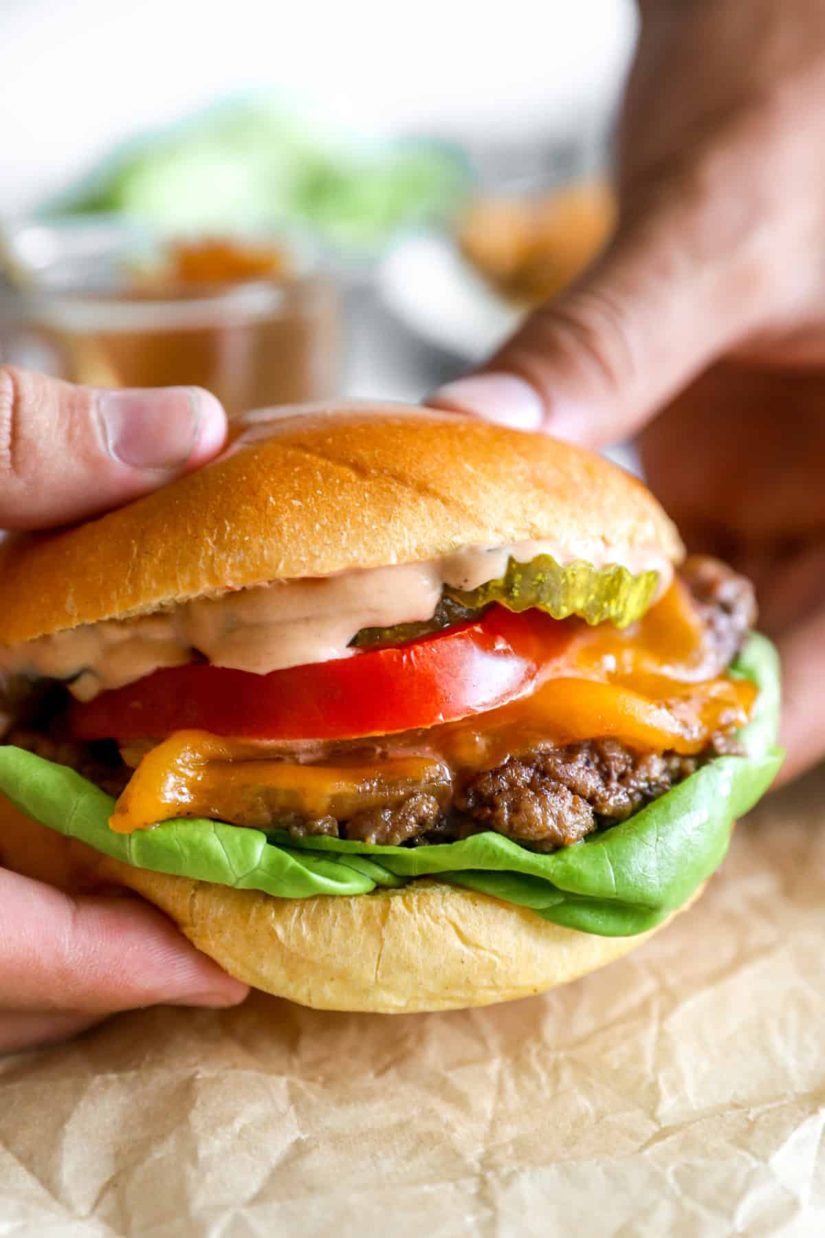 Two hands holding a smash burger with lettuce, tomato, pickles, and burger sauce.