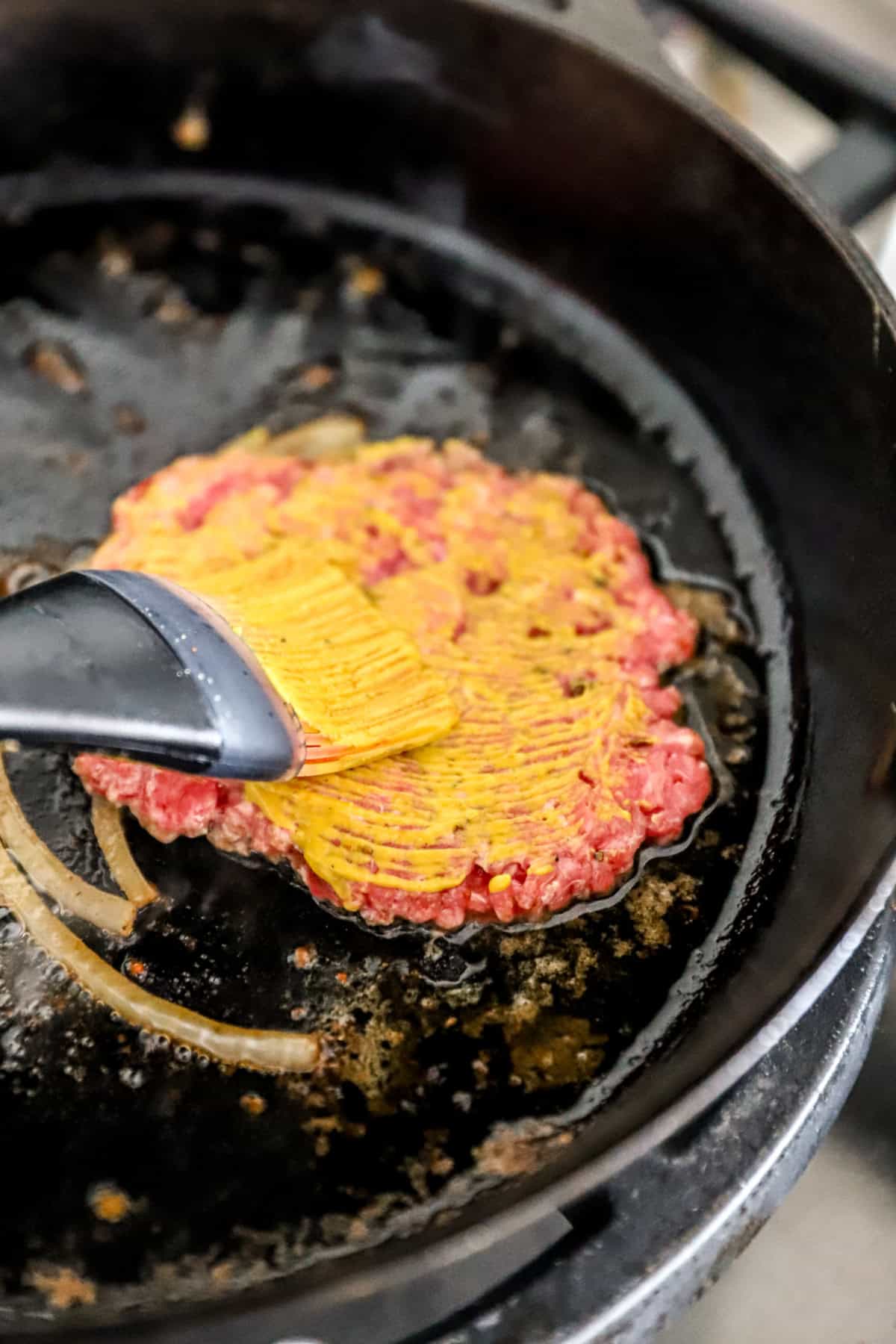 Mustard being brushed over smash burgers in a skillet.