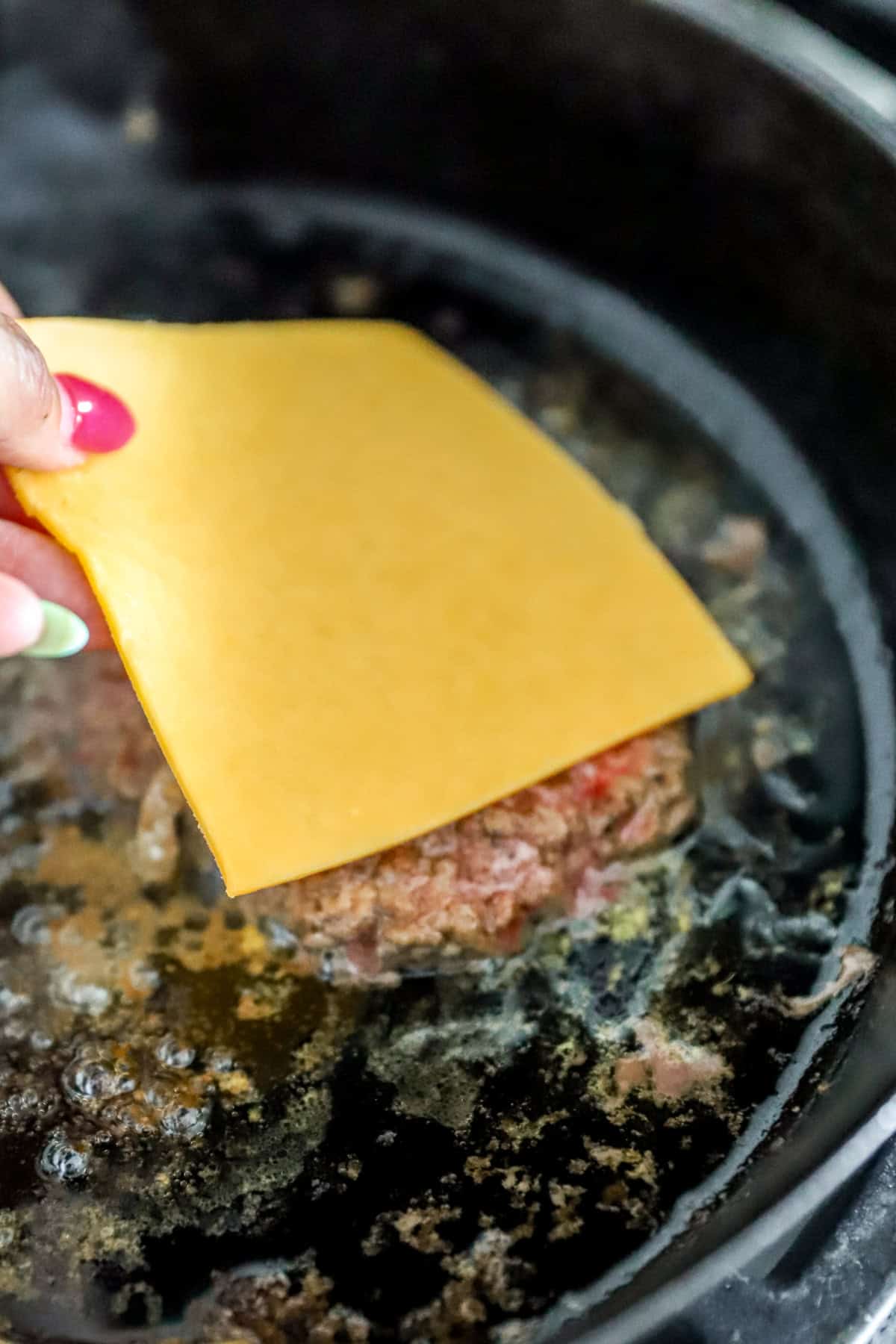 Cheese slices being placed over smash burgers cooking in a skillet. 