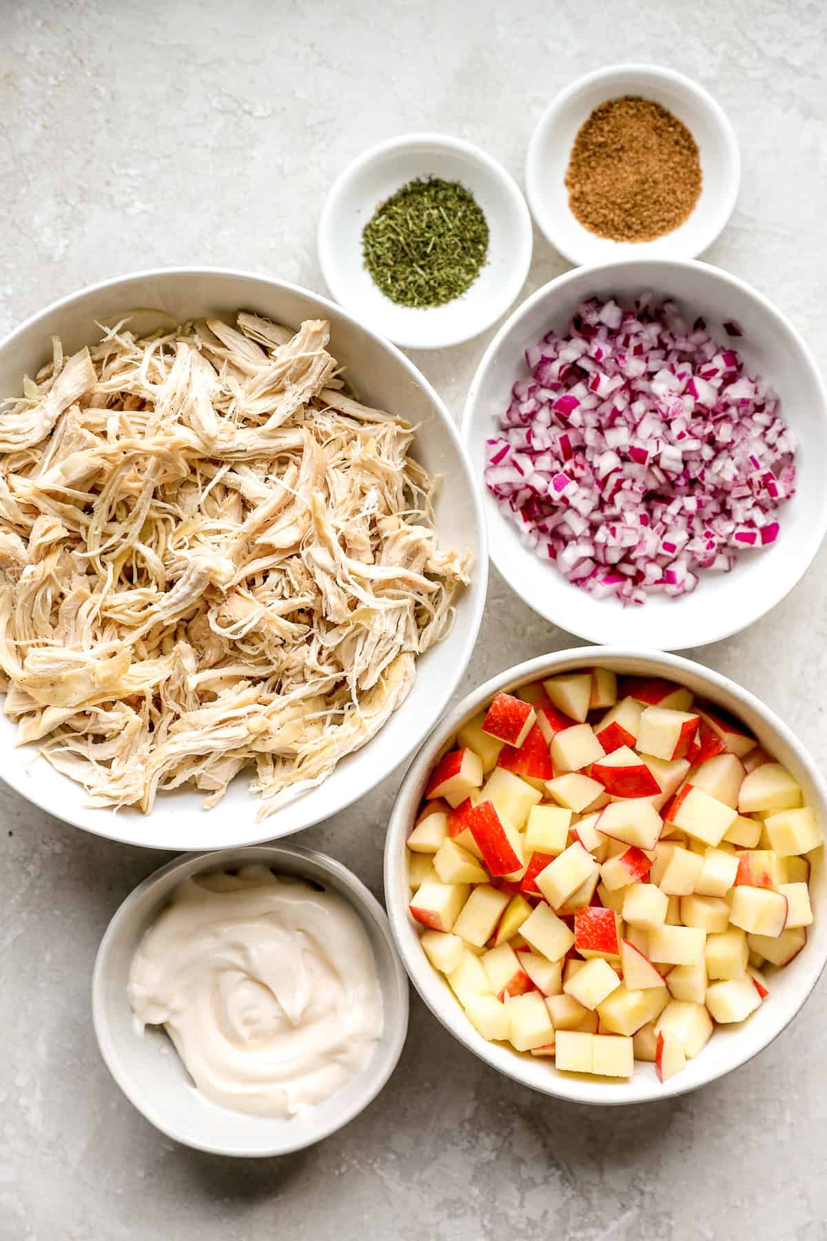 Ingredientes de ensalada de pollo de manzana en varios tazones: pollo rallado, manzanas, condimentos, cebolla y salsa.