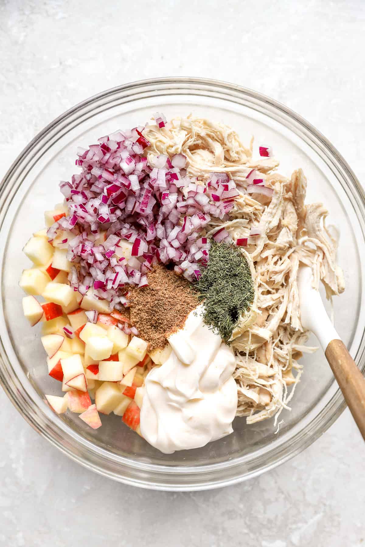 Ingredientes de ensalada de pollo de manzana en un tazón de vidrio transparente con una espátula blanca.