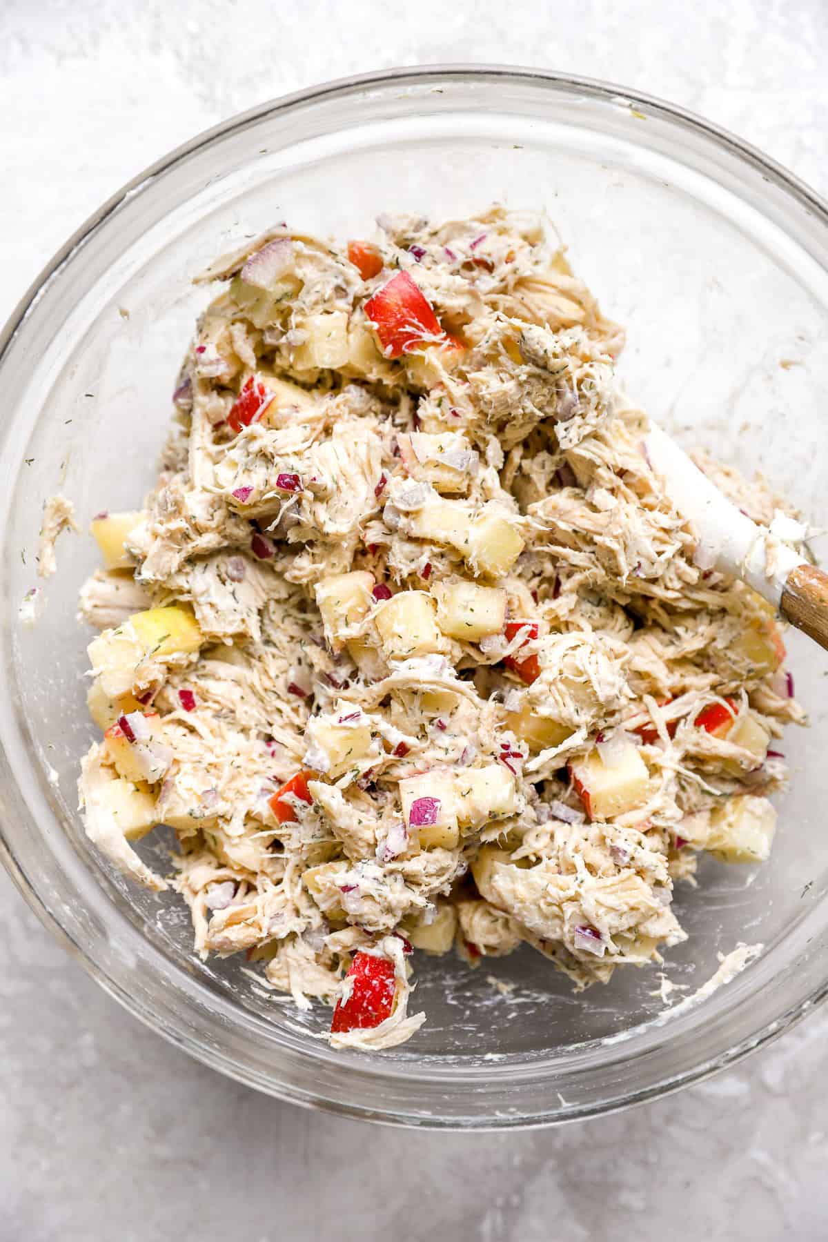 Ingredientes de ensalada de pollo de eneldo de manzana completamente mezclados en un tazón de vidrio con una espátula blanca y de madera.