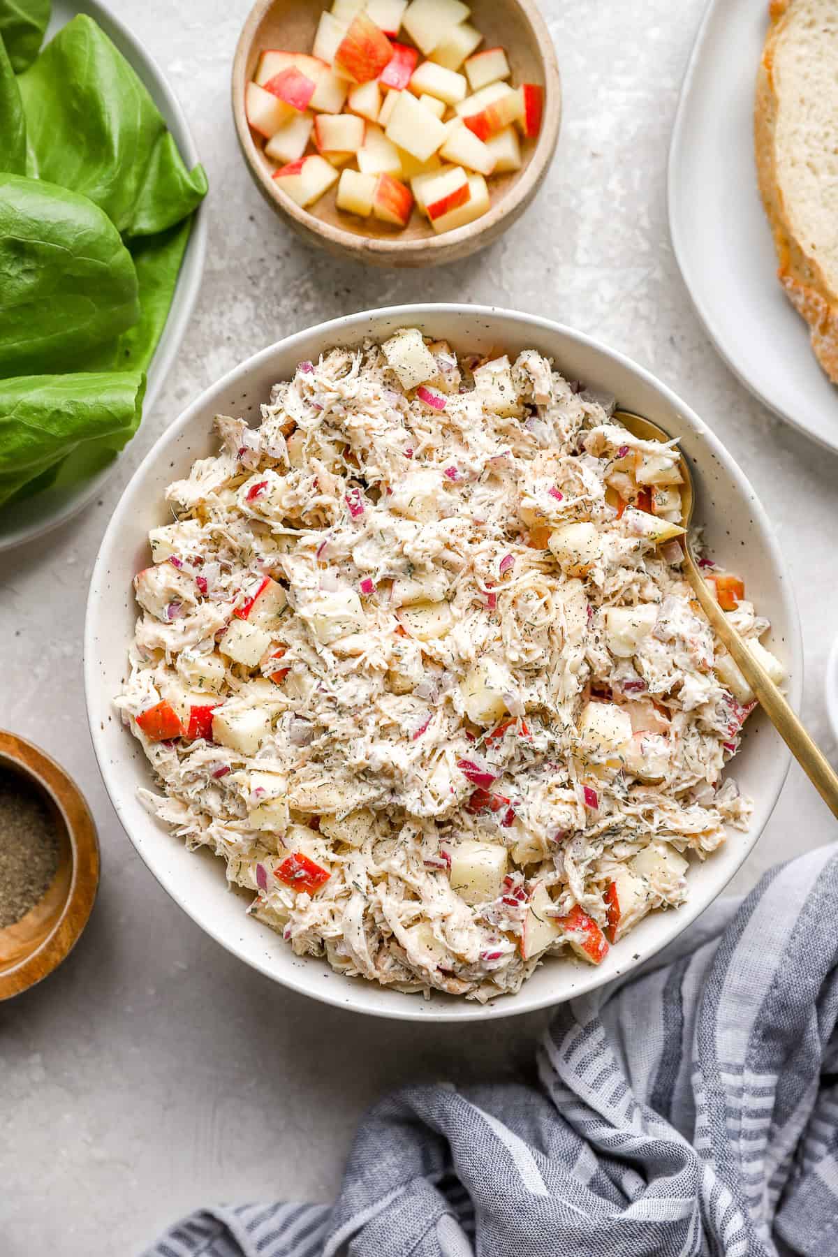Ensalada de pollo de eneldo de manzana en un tazón blanco con una cuchara para servir de madera y en el mismo mostrador hay mantequilla de lechuga y especias.