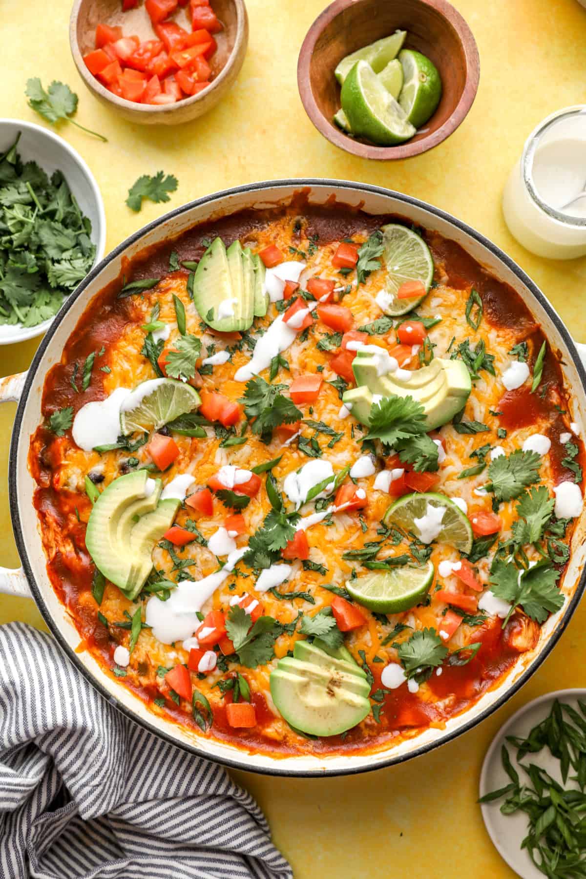 Overhead image of a skillet full of chicken skillet enchiladas topped with lime wedges, avocado slices, and cilantro. 