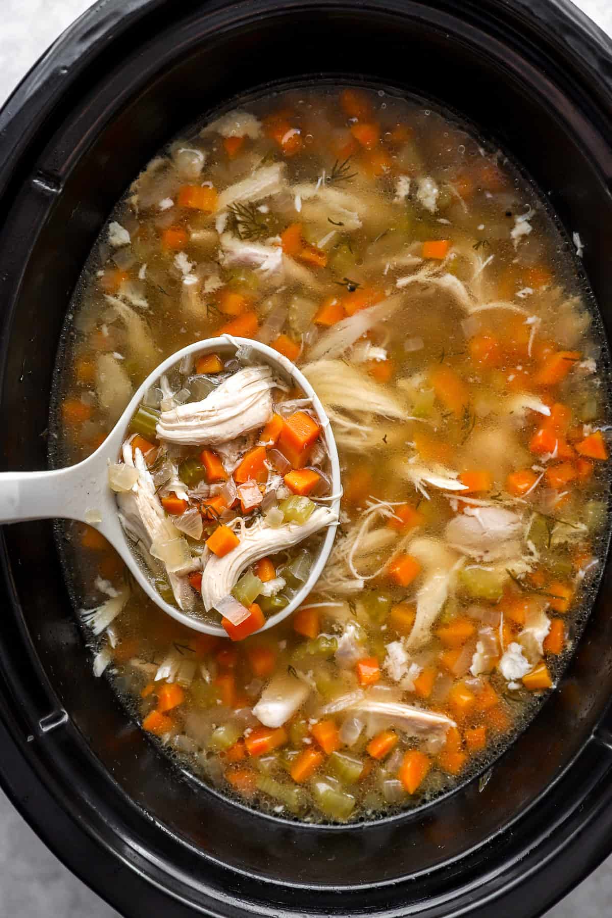 A ladle lifting a scoop of chicken soup from a slow cooker.