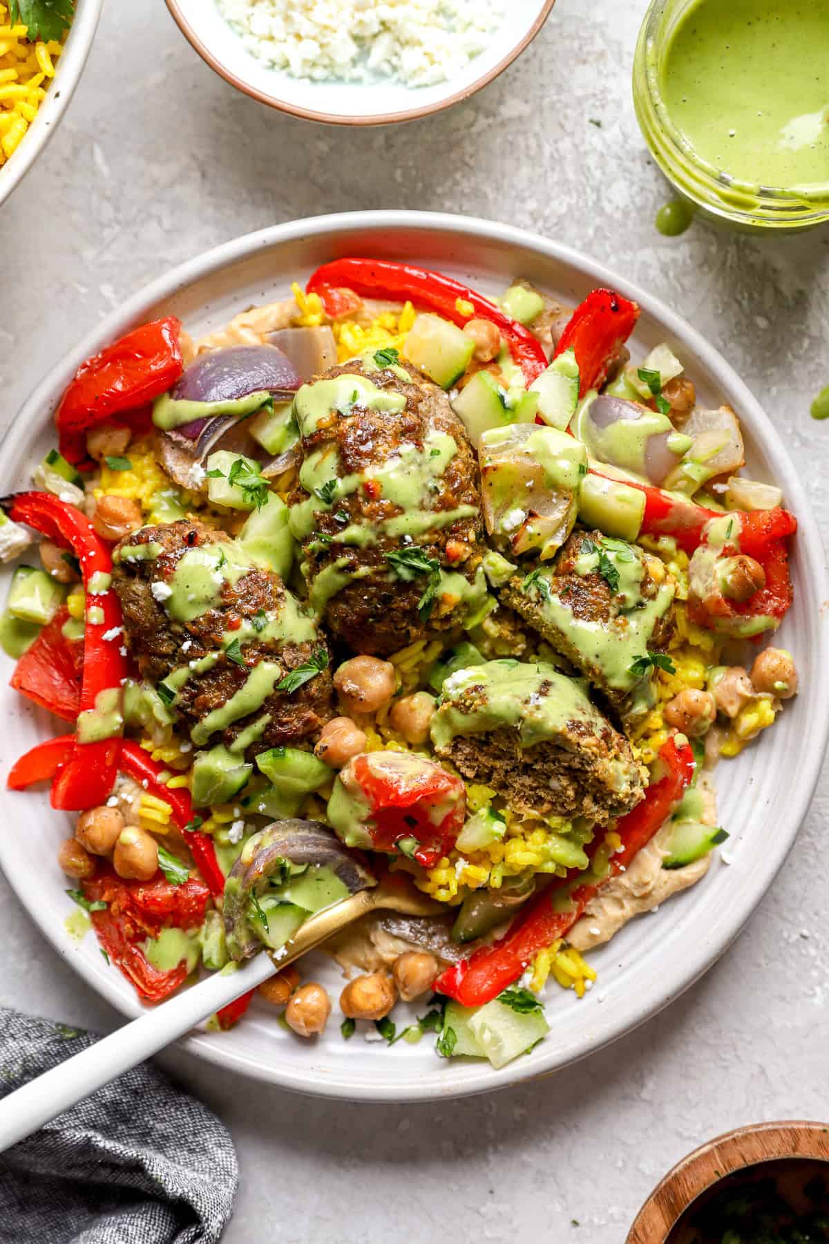 A fork on a plate with kofta meatballs, veggies, chickpeas, turmeric rice, and green tahini.