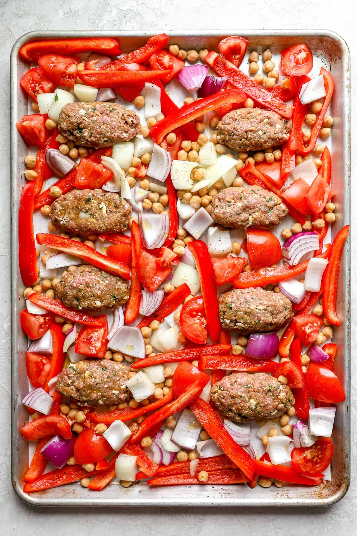 Raw kofta meatballs on a baking sheet with veggies and chickpeas. 