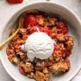 A bowl of strawberry crisp topped with vanilla ice cream with a spoon on the side.